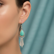 Charger l'image dans la galerie, A pair of silver dangle earrings with teardrop-shaped turquoise stones and dangling fringe, displayed on a textured light blue fabric background.
