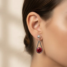 Load image into Gallery viewer, Silver dangle earrings with a teardrop-shaped design, featuring a polished silver post and a drop composed of a red faceted crystal and a silver disc with a red band, displayed on a textured light blue and white surface.
